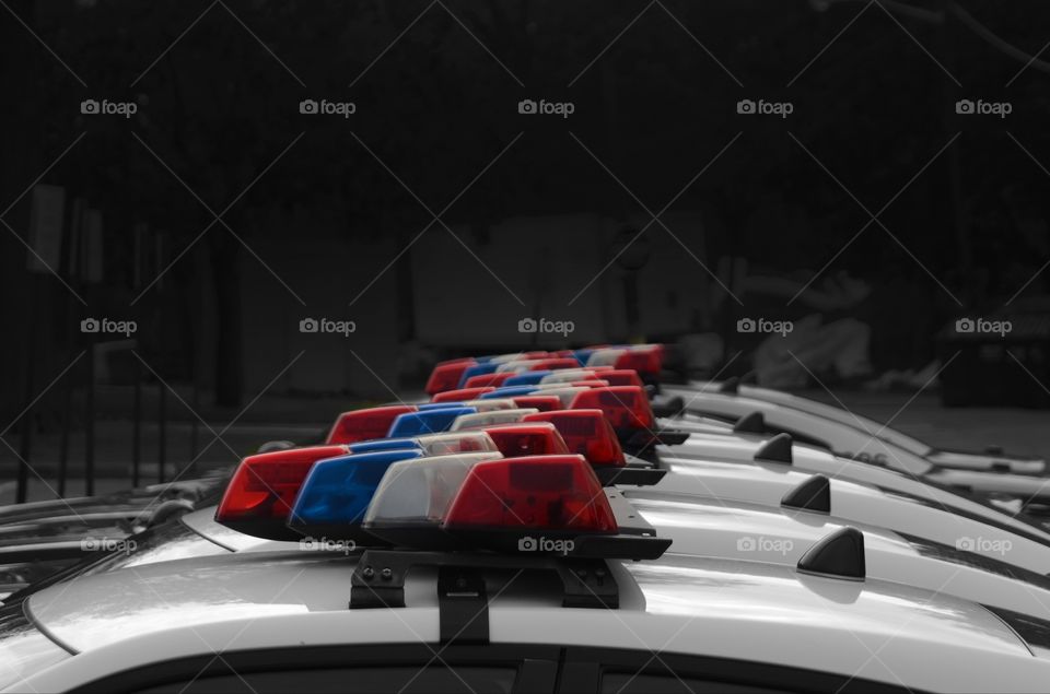 A fleet of black and white police cars with the red, white, and blue berry lights atop are lined up on a residential street.