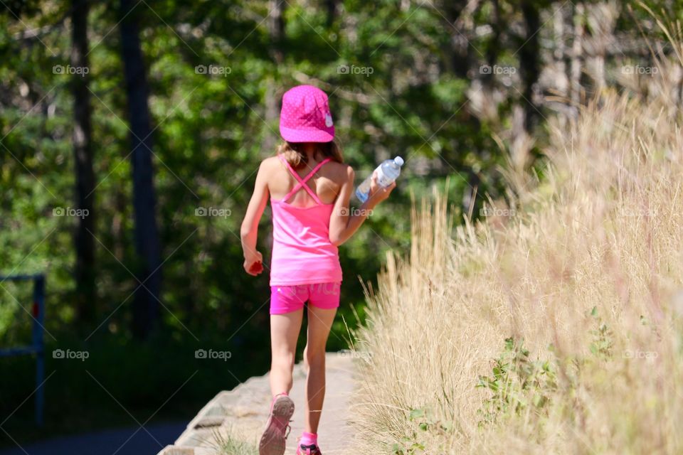 Young girl child dressed in pink baseball hat, top and shorts and holding water bottle walking across ledge in park