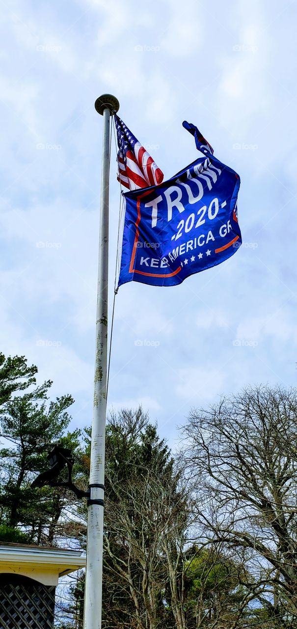 Trump 2020 flag flying straight out on flagpole in the wind under the American Flag. These flags of represent freedom as they fly against a blue sky.