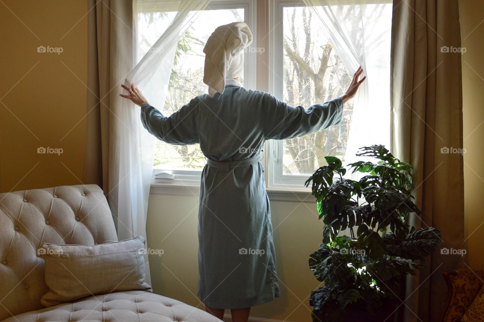 Woman from behind wearing a robe and toweled hair opening the curtains of a bedroom window in the morning