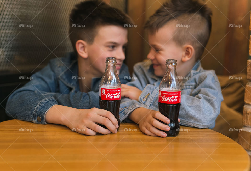 two lovely brothers, dressed in denim clothes, with beautiful hairstyles, are sitting in a cafe and drinking Coca-Cola. communicate, laugh, smile. two guys, two friends