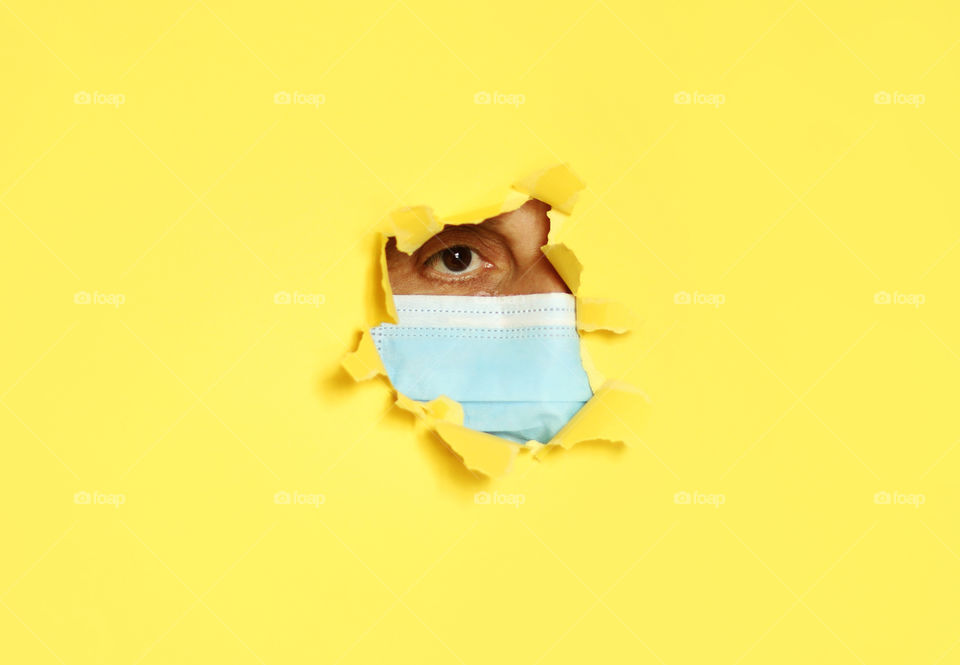 Isolation. Woman's face with protective mask in the hole of yellow wall paper