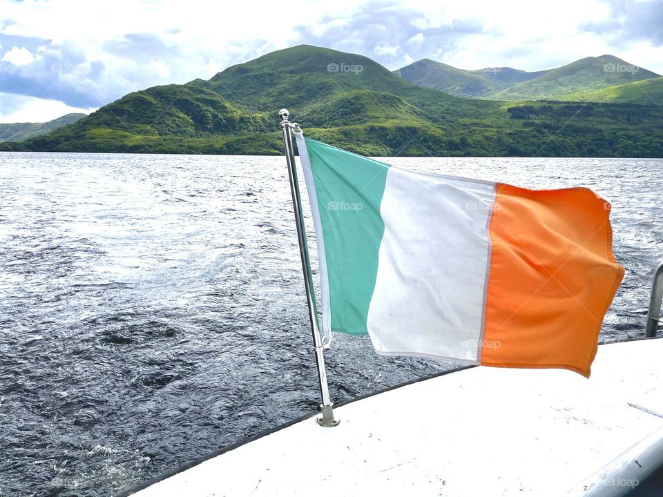 An Irish flag at the stern of a boat on Killarney Lake waves proudly in the strong wind against the lush green hills of the shore.