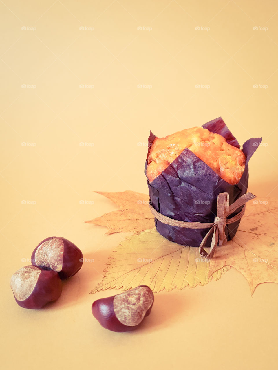 Autumn still life with pastries. Muffin in brown baking paper with chestnuts on autumn leaves on a plain beige background. Beginning of autumn