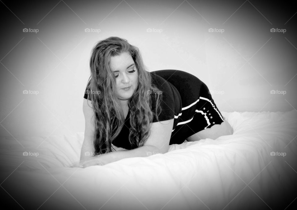 Soft, plus size woman posing on white bed, looking down. She has long hair and wears a black dress. Black and white photo.