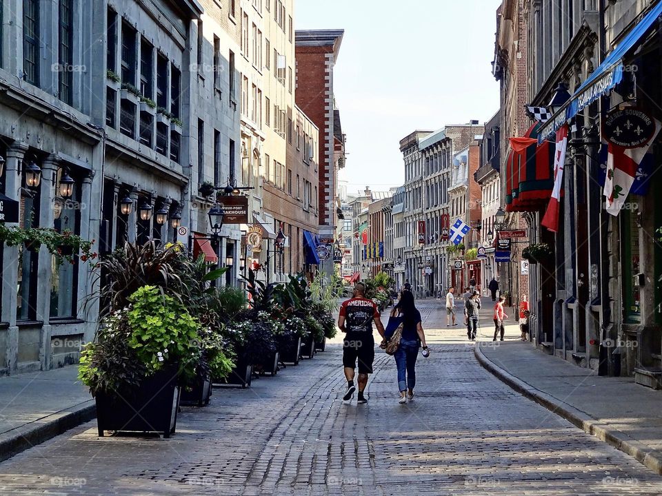 Old Montreal , cobblestone street . Couple walking down centre of street beautiful architecture on both sides of street , plants line one side of the street