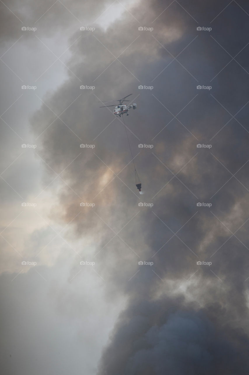 Helicopter of State Emergency Service is bringing water in the bucket. Smoke from the fire in the background