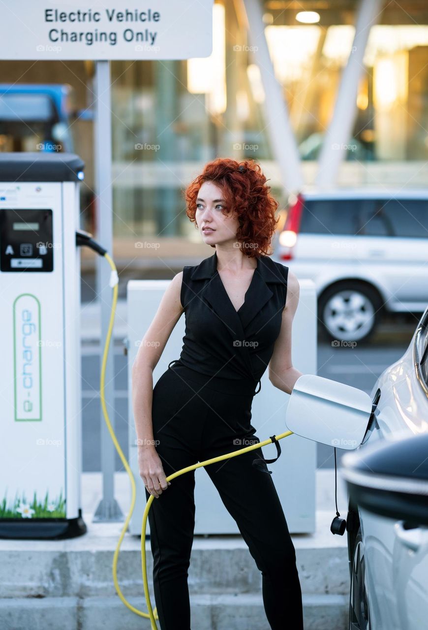 A woman charging electric car at charging station. Full body portrait of red hair woman in black suit