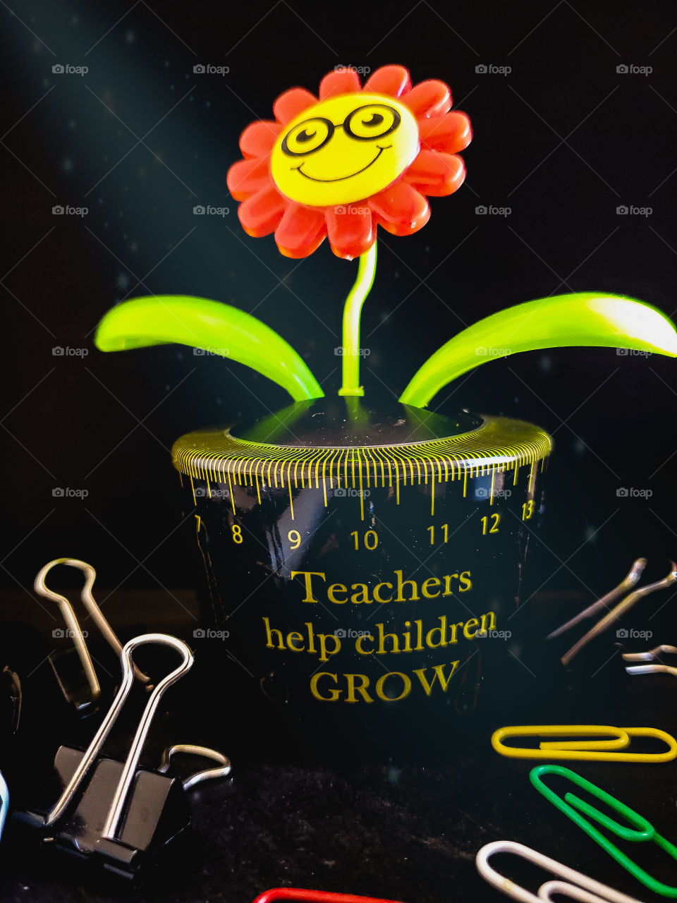 Teacher Appreciation. Still life of a solar plant with magic light shining down. The message on the solar plant says teachers help children grow and it is surrounded by paper clips and binder clips on a black background.