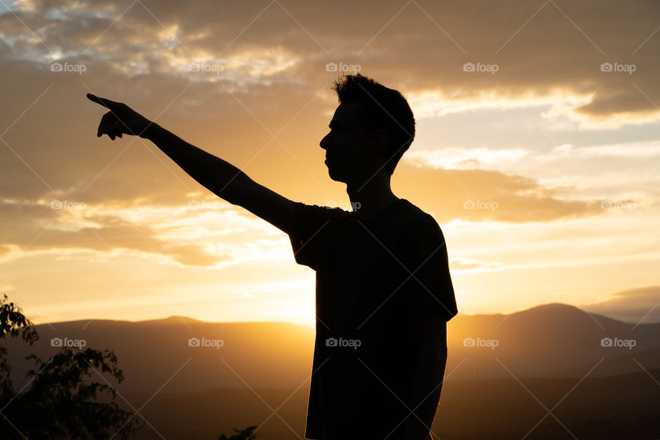 A man silhouette points to the side after hiking up a mountain during sunset 