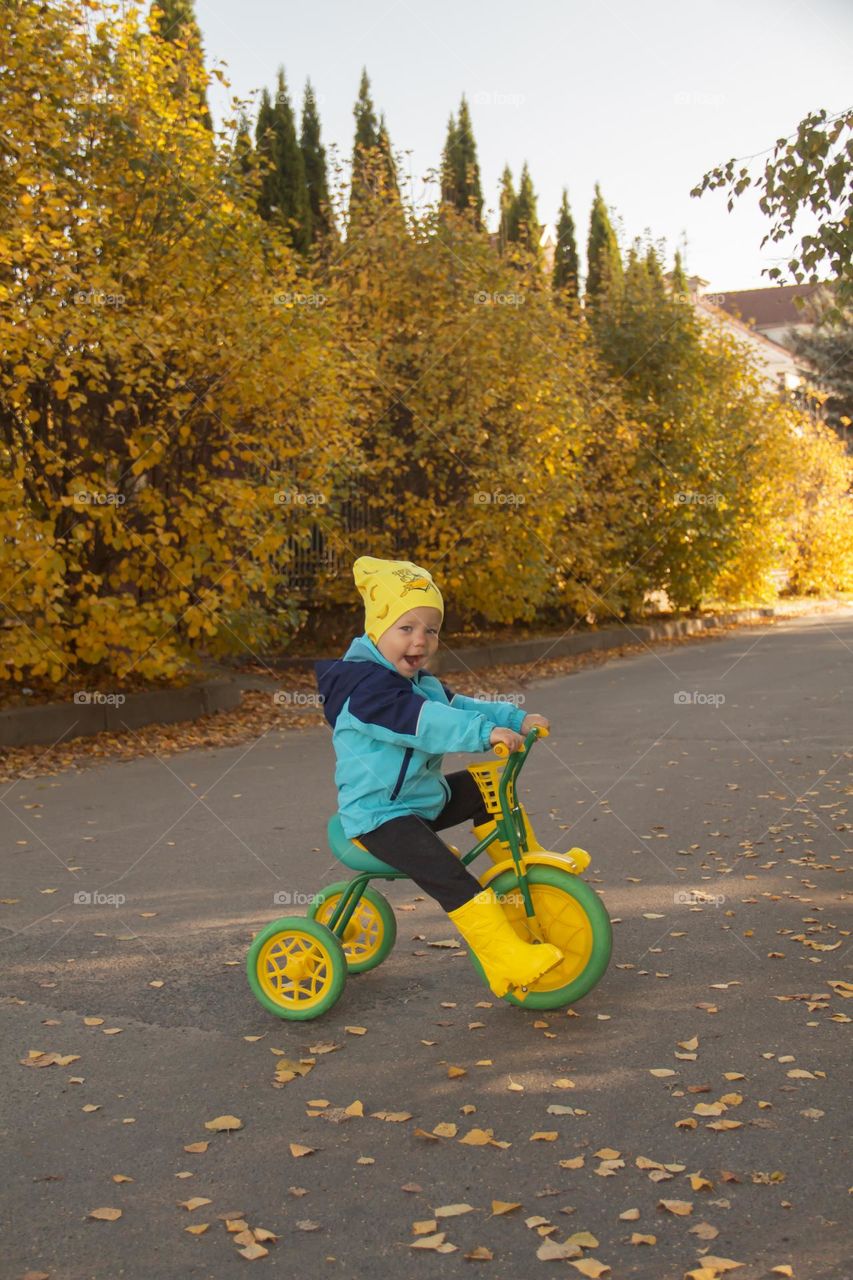 a little boy in a yellow hat and yellow boots rides a bike in autumn