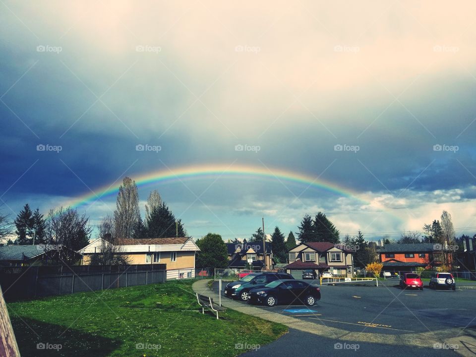 Rainbow in a cloudy sky, light rays through clouds. Green grass and a parking lot.