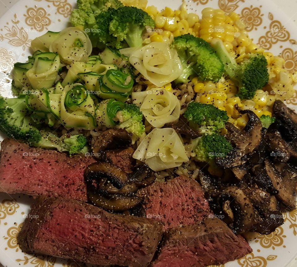 Sliced beef tenderloin with mushrooms, steamed broccoli, strips of zucchini and white carrots, folded into floral shapes, fresh corn cut from the cob, and served on a bed on garlic and herb quinoa and brown rice.
