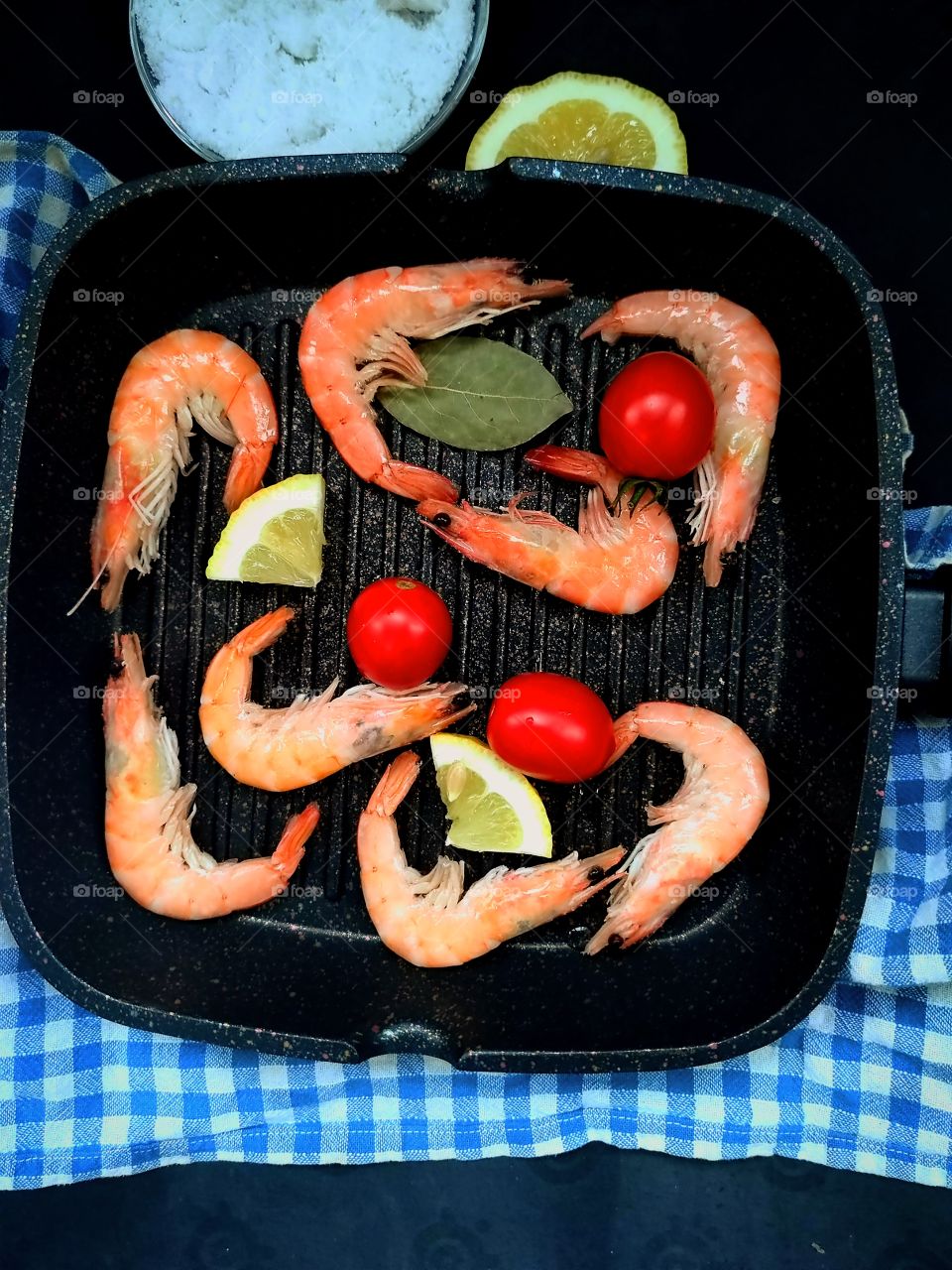 Black skillet containing shrimp, cherry tomatoes, lemon wedges and bay leaf.  Salt and half lemon are lying next to a frying pan. Top view. Black background
