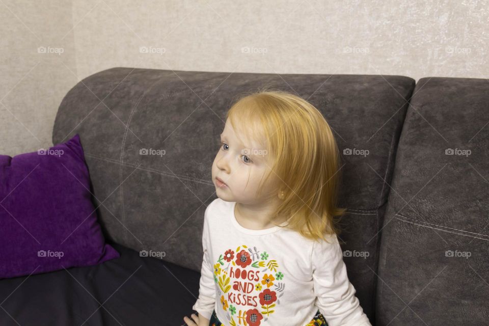 A small, red-haired girl is laughing at home on a gray sofa with purple pillows and a yellow background