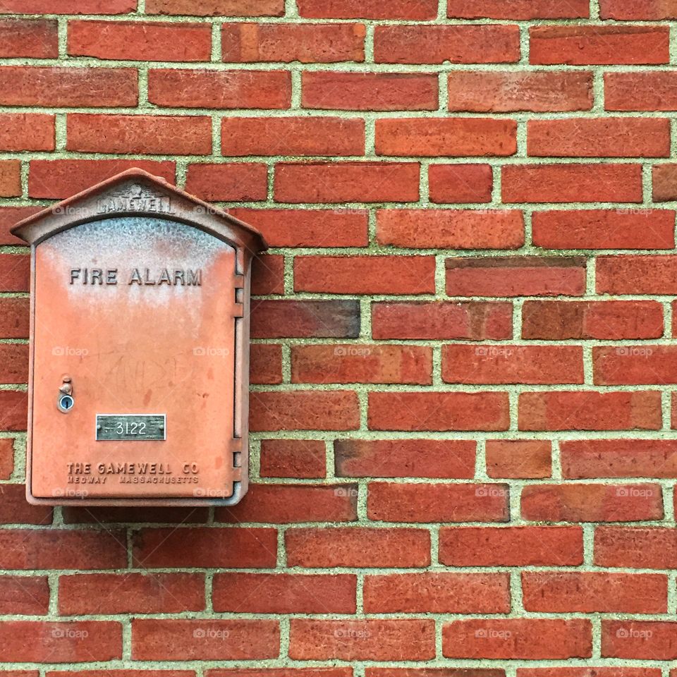 A fire alarm box mounted on an exterior brick wall. The principal color of the image is reddish brown.