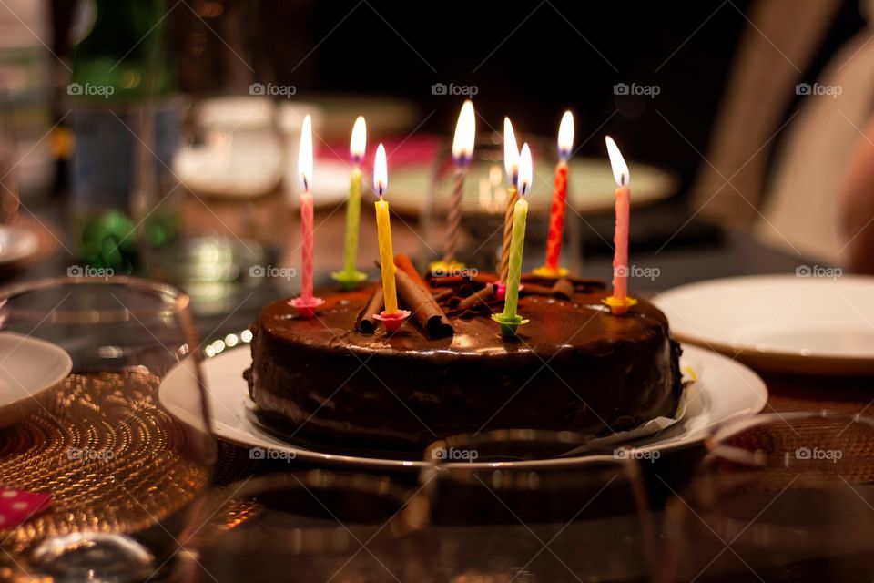 a portrait of a chocolate birthday cake with burning candles on it on a festive dining table. the lit candles all have different colors.