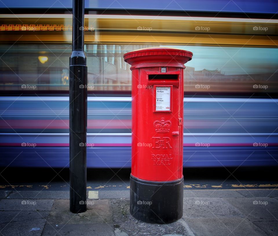 A bright red Royal Mail post box on the city streets with a motion blurred bus in the background creating a colourful abstract image.