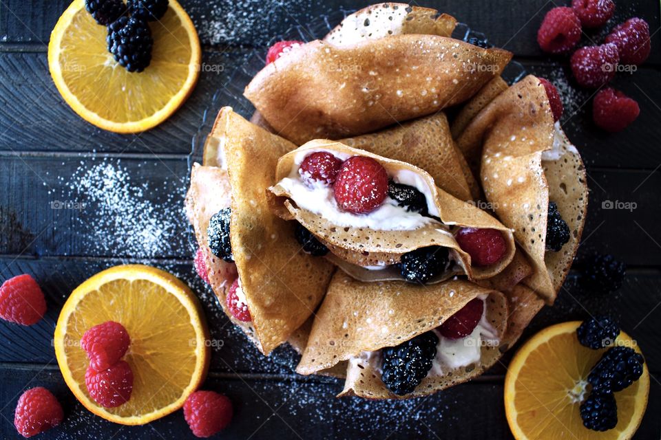 A plate of homemade cream and berry filled crepes on a dark widen board with orange slices, raspberries and blackberries around it