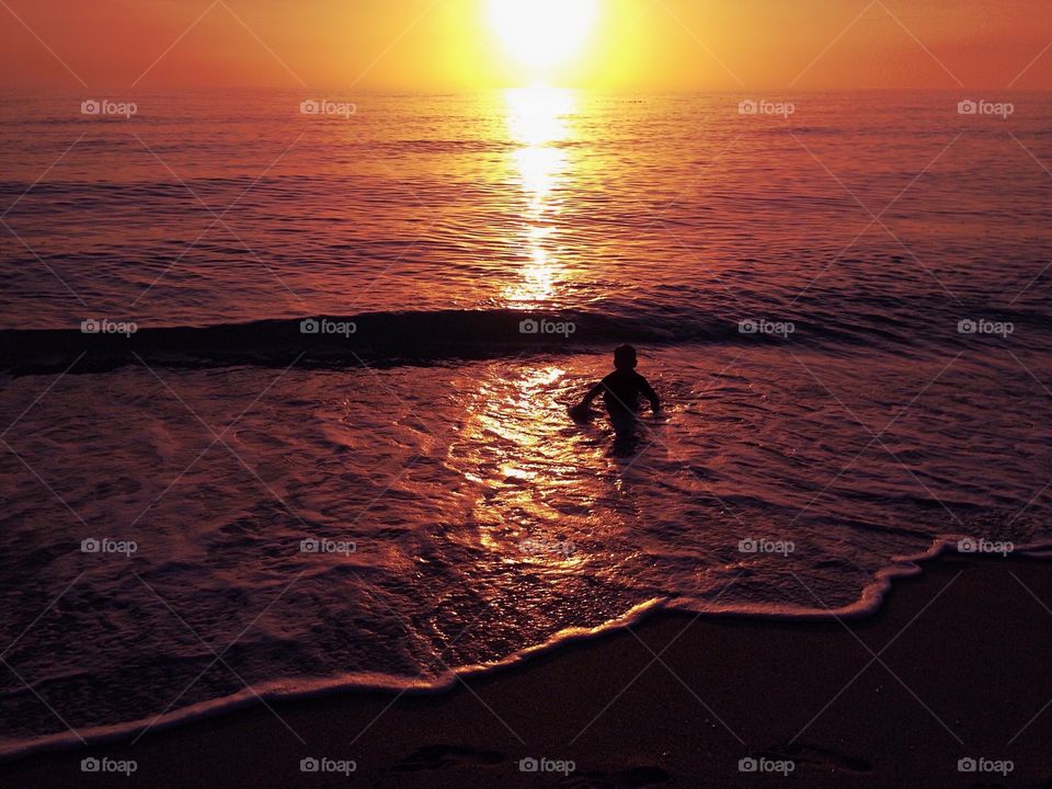 Small boy’s silhouette in the ocean surf surrounded by an intense amber sunset.