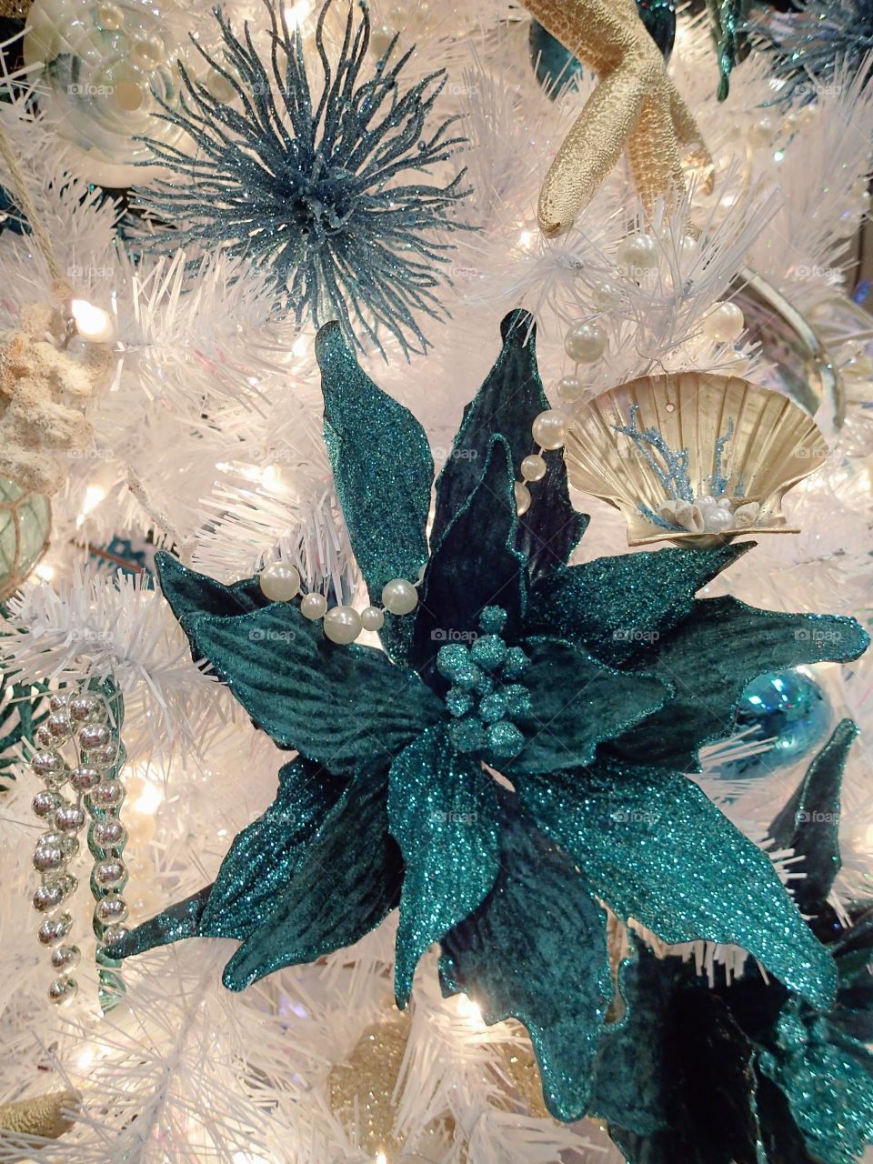 Bright glittering ornamental turquoise flower hanging from a white Christmas tree. 