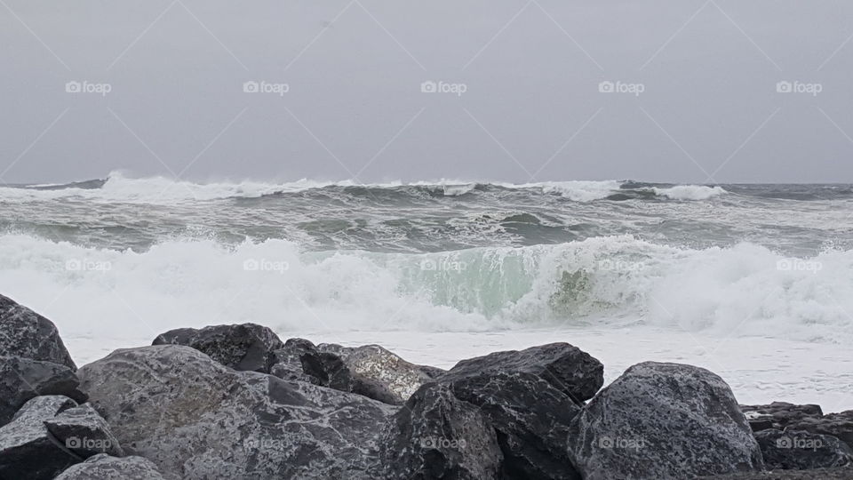 Waves crashing in to rocky shore, rolling towards viewer. Grey sky with choppy water.