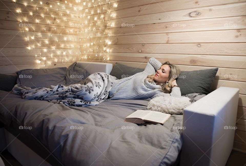 Young beautiful woman relaxing in cozy bed with book open on christmas lights bokeh background