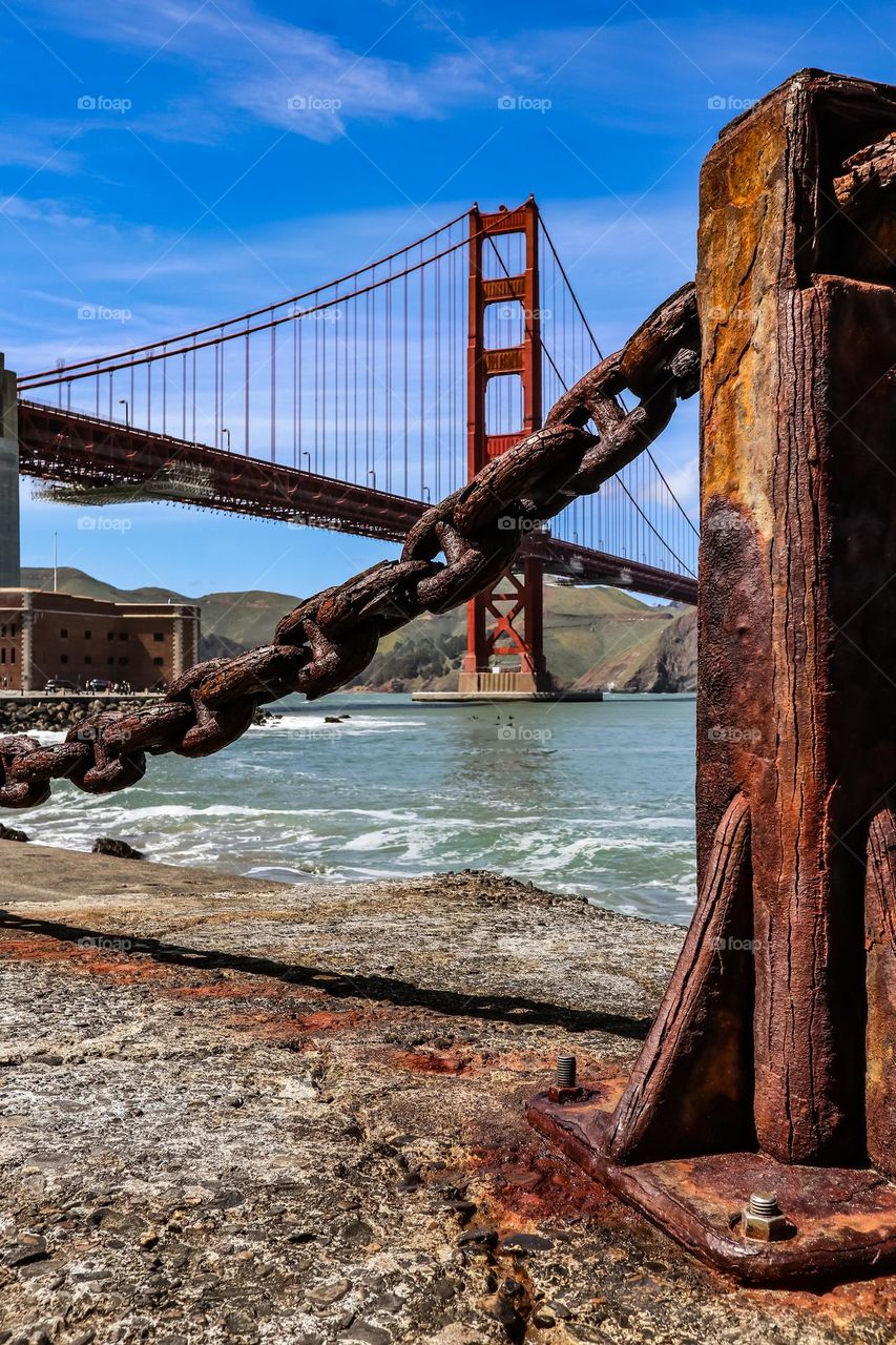 Ground level view of the Golden Gate Bridge from Fort Point in San Francisco California through the rusted iron chain link guardrails and posts that have oxidized over time appearing almost wood grain like.