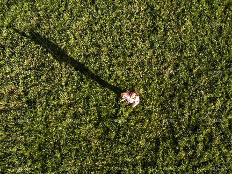 A little girl standing on a field, a drone is flying above her taking her picture as she looks up and waves