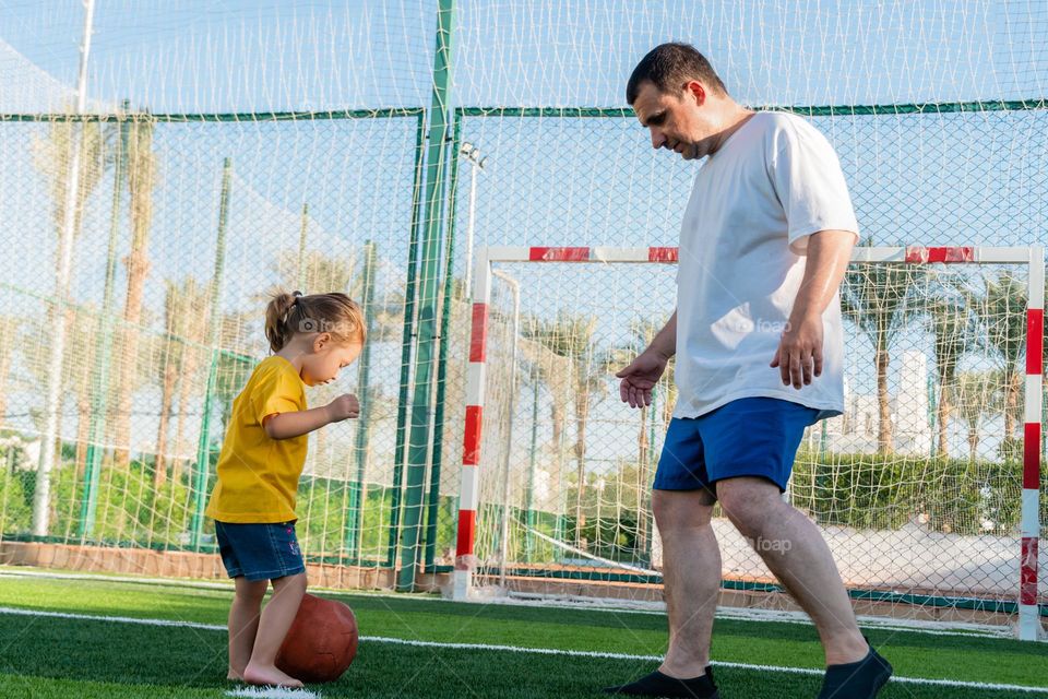 Full body positive man playing soccer with little daughter kicking ball on green lawn. Father's Day concept. Joy of life.