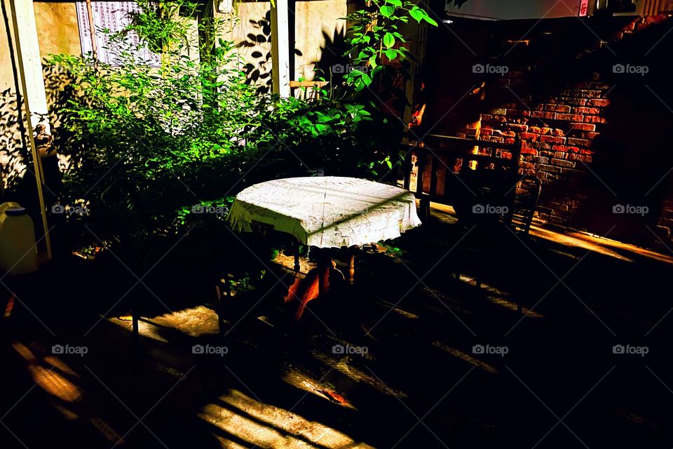 A table at night with a white linen cloth on top. There is greenery in the background and a brick wall in the back. 