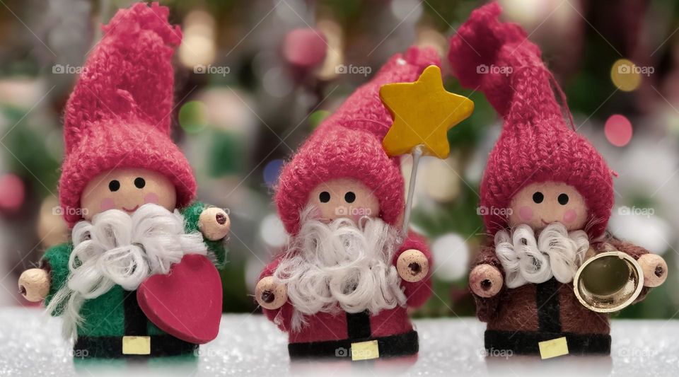 3 tiny Christmas folk made of wood, with magenta, knitted hats against blurred background of lights & decoration 