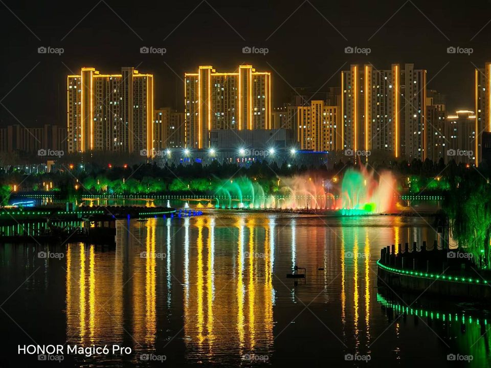 This photo showcases a beautiful night scene with tall buildings, reflections and fountains illuminated by colorful lights. It exudes endless beauty through the depiction of light and is incredibly stunning.Tt was taken in China and is intended for c