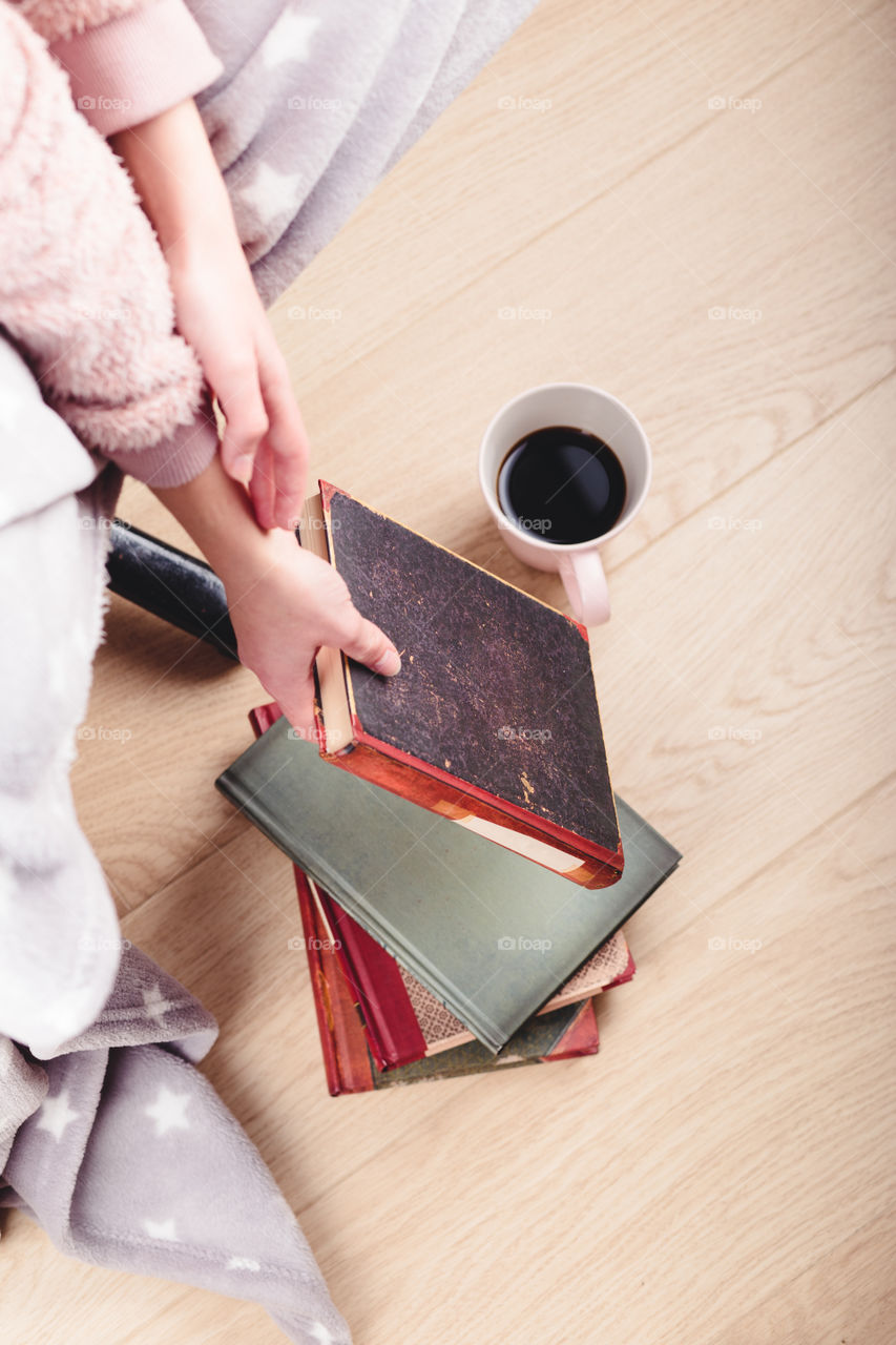 Girl enjoying the reading a book and drinking coffee at home. Young woman sitting on a chair, wrapped in blanket, holding book, relaxing at home. Portrait orientation. View from above