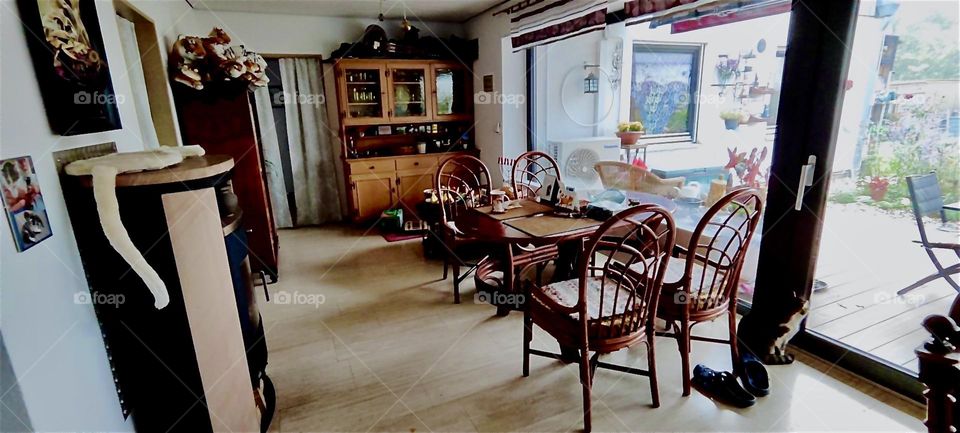 This is a larger view of the third section of the L shaped living room in the house in „Bavaria“, Germany showing the entrance and a wooden wardrobe with glass doors. All tables have a view of the garden. 2023. Hypnotic Productions