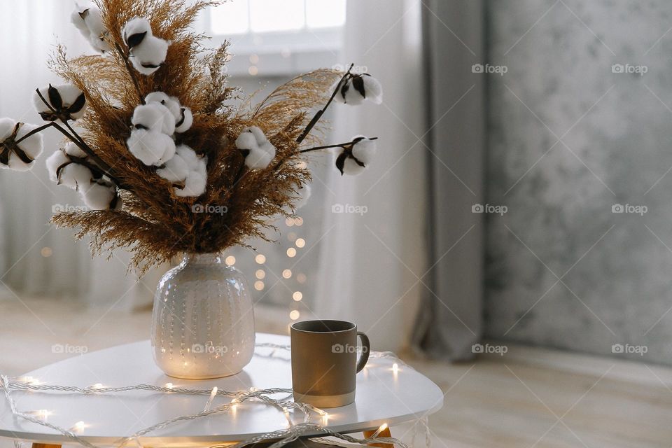 Cup of coffee and vase with cotton flower on white table in room