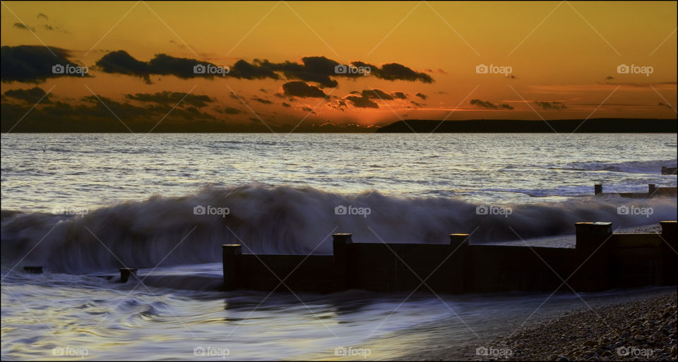 A wave rolls of the wooden groaned in front of a deep orange sunset - Bexhill beach, U.K.