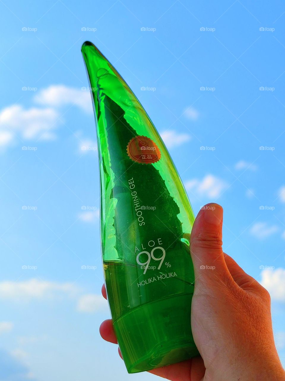 A woman's hand holds a green bottle with a white inscription: "HOLIKA HOLIKA ALOE 99" against a blue sky and white clouds. Holika Holika Aloe 99% Soothing Gel soothing and moisturizing with aloe