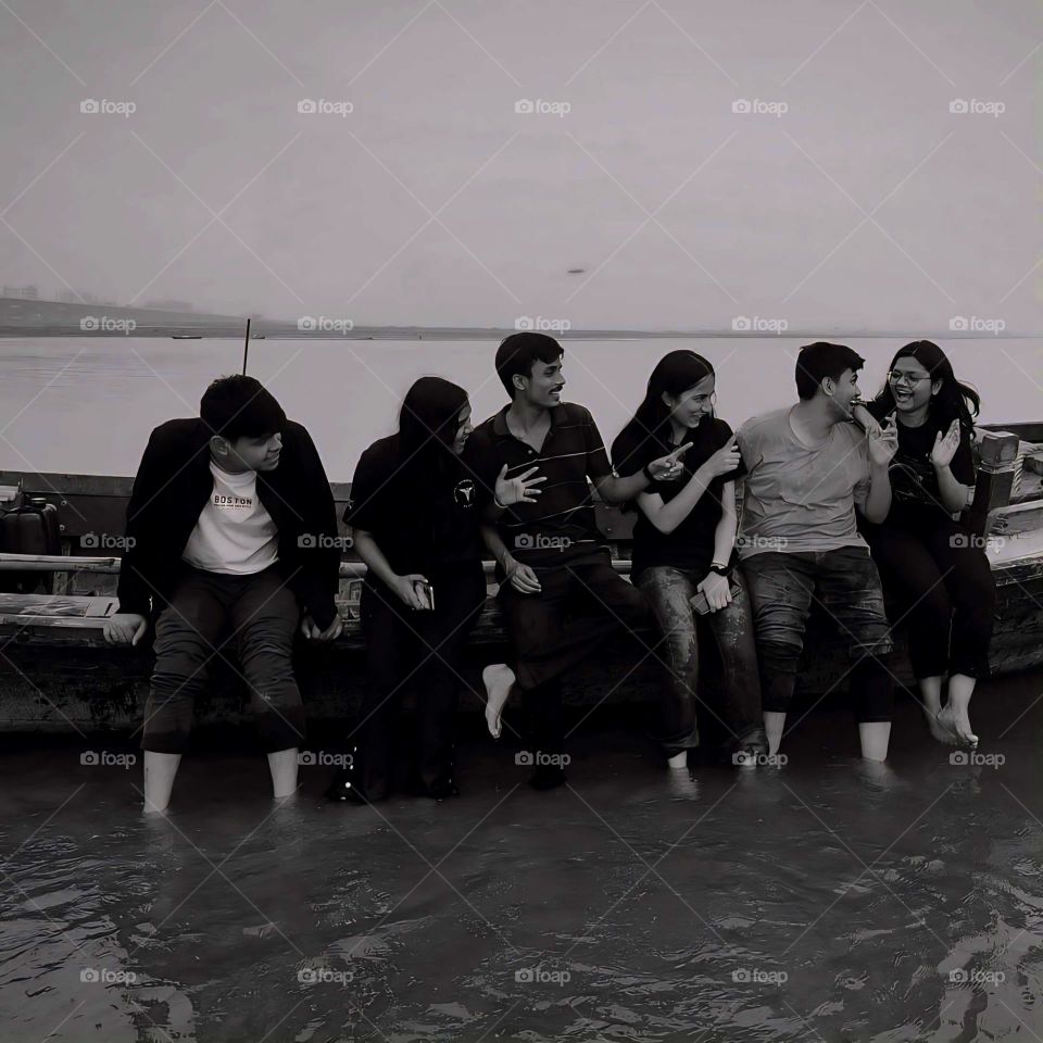 The monochrome palette adds a timeless quality, highlighting the friends' emotions and expressions. Their casual, barefoot postures emphasize relaxation and spontaneity, capturing a beautiful, carefree moment in a tranquil setting.