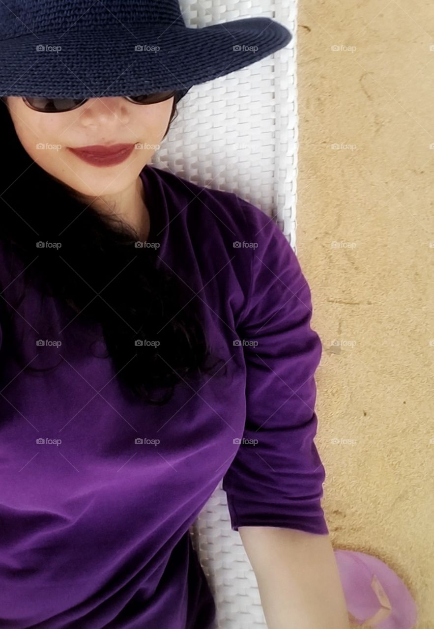 A selfie with my long hair relaxing on a white hammock wearing a sunglasses, a blue hat and purple shirt with my thong sleeper on the sand.