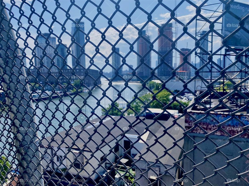 Through the mesh fence on top of the “Pulaski Bridge” at “Newtown Creek” an entire panorama of “New York City” can be seen from the “East River” to LIC, Queens and “Manhattan” and to the left “Greenpoint”, Bklyn. 2023. Hypnotic Productions