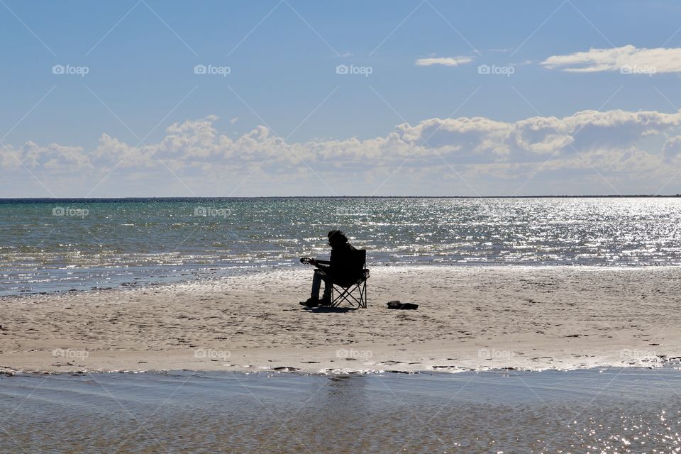 Stranded musician sitting in chair at ocean at low tide surrounded by water playing guitar and singing, golden hour glistening 