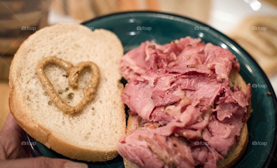 Holding a plate with famous Montreal Smoked Meat on Rye Bread with mustard heart and loaf of rye bread on table in background 