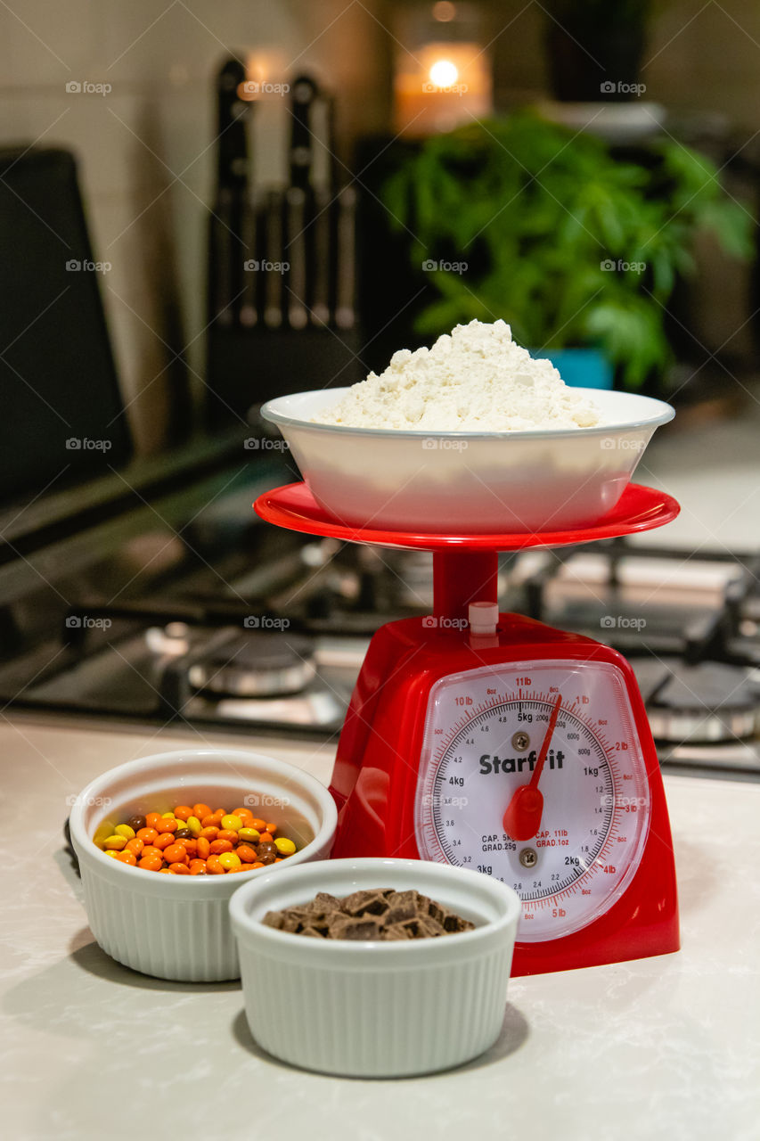 Flour in a bowl being weighed and measured on a food scale, with chocolate chunks and peanut butter pieces in small dishes for cookies.