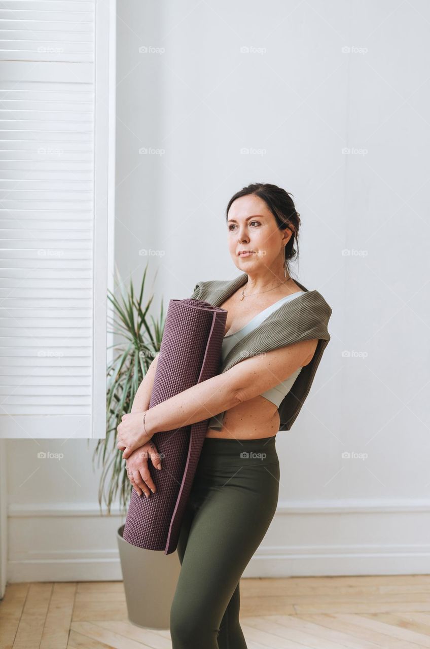 Attractive middle aged brunette woman in sportswear with yoga mat in the light studio