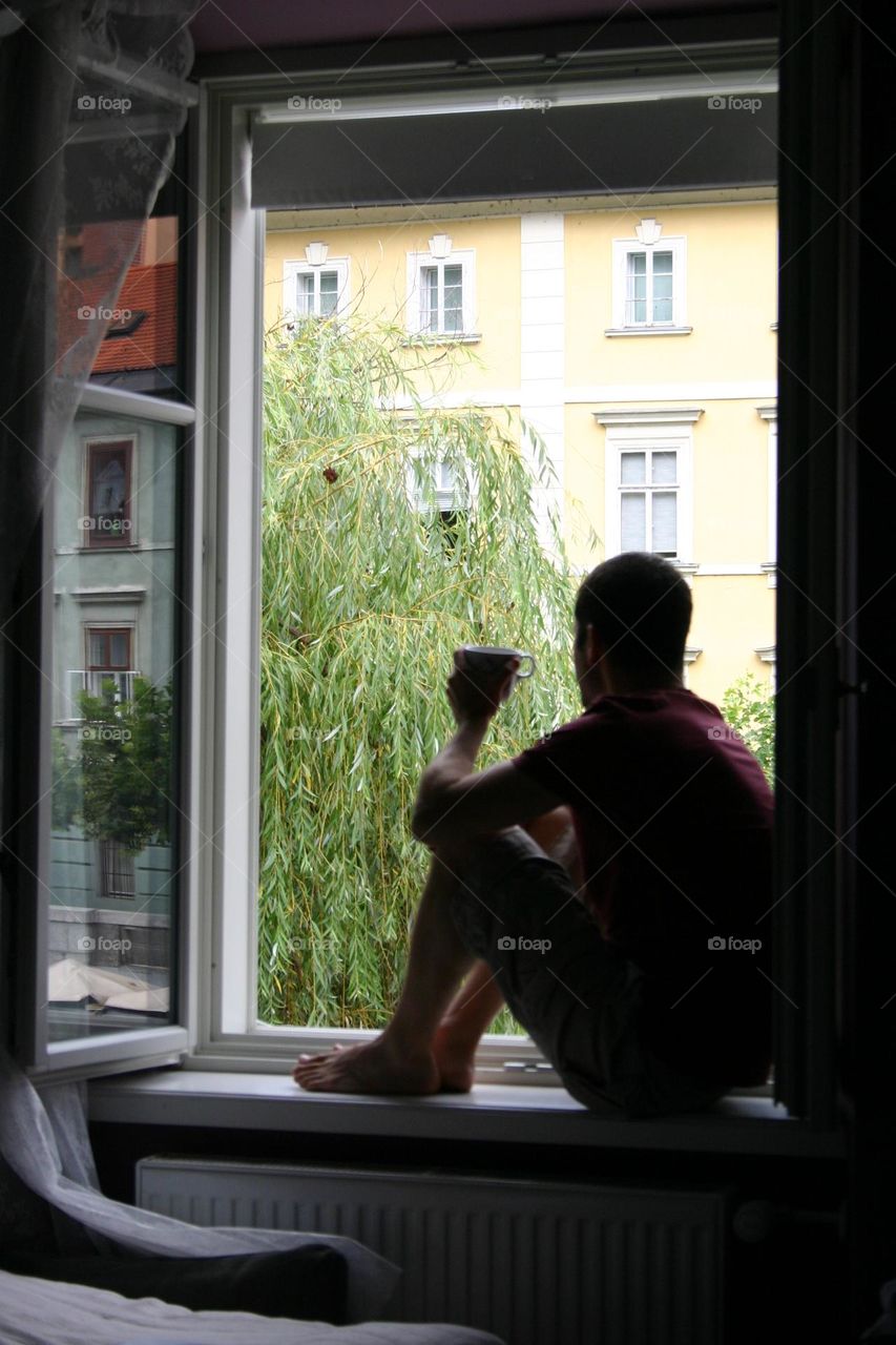 Man sit on the window looking outside with a cup of coffee in his hand