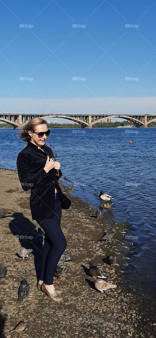 девушка в темных очках на берегу реки, в воде плавают утки, на фоне городского моста