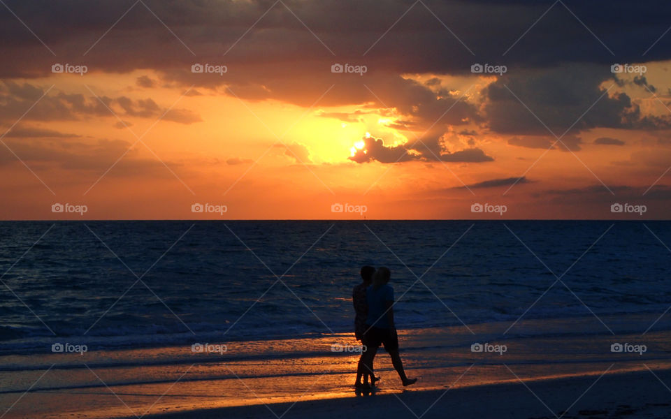 Silhouette of Couple strolling by seashore at sunset under fiery golden yellow red cloudy skies horizon in background