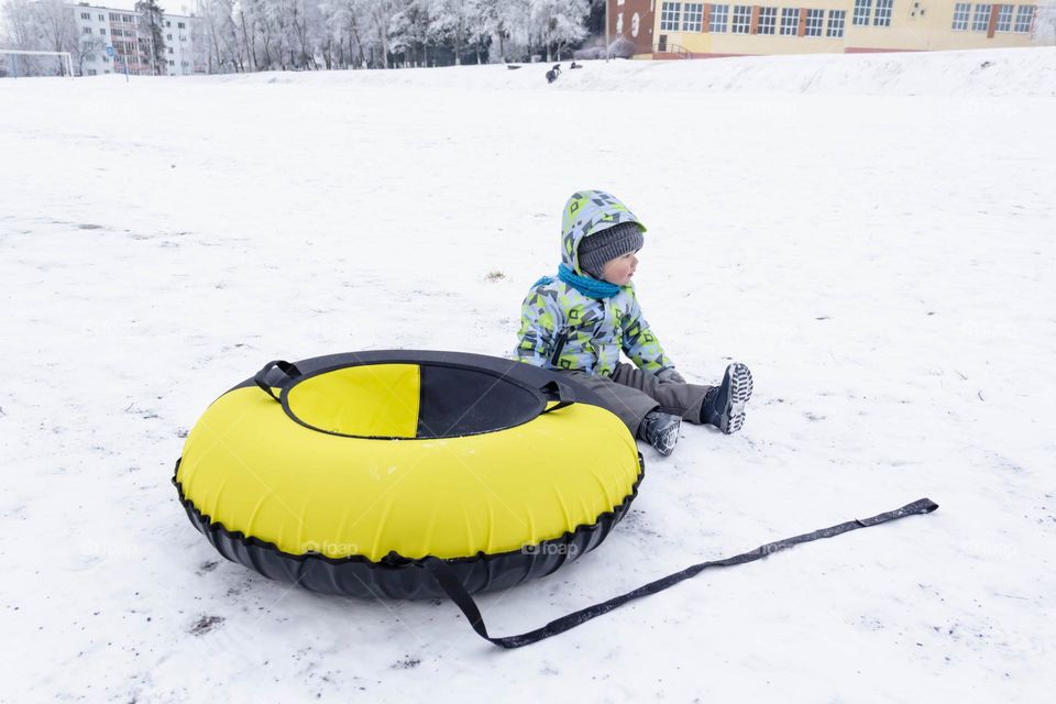 A small, carefree boy walks in the white snow in winter and rides a tubing in the park, near trees in the snow.