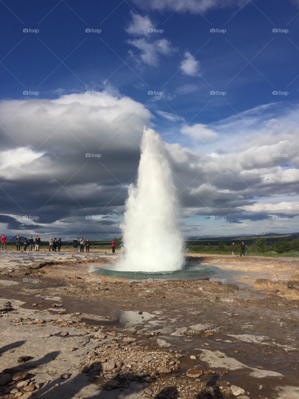 Geyser erupts from the ground! Blue skies with white clouds, green fields in the background. 